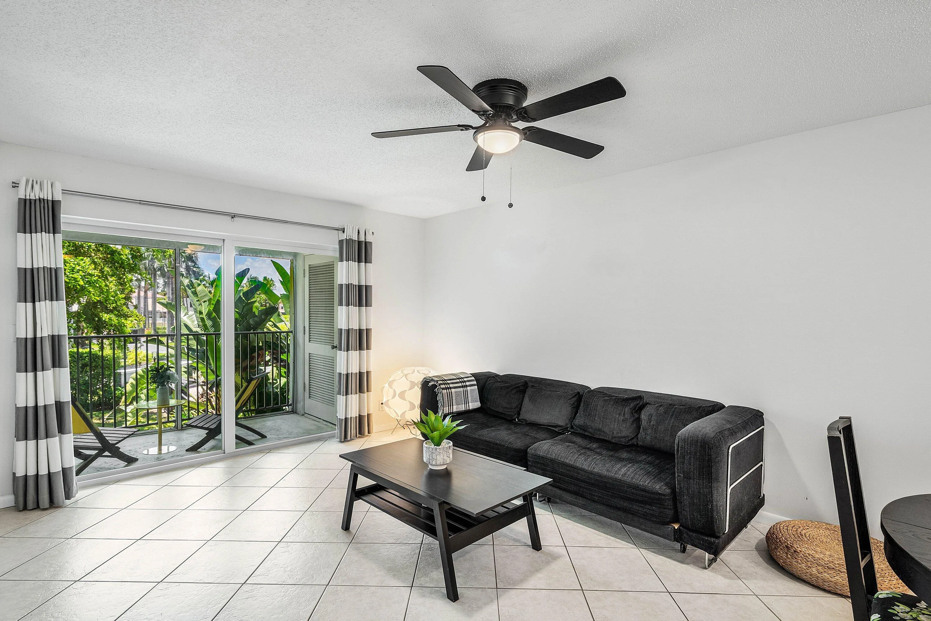Spacious living area featuring light-toned square tile flooring, a dark-toned ceiling fan with integrated lighting, and a private balcony with a dark-toned railing