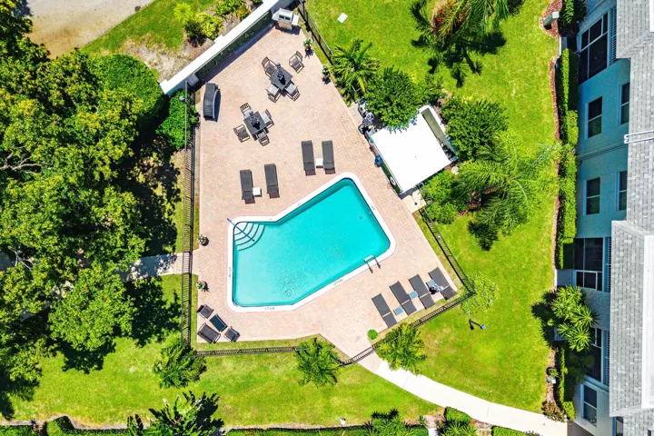 Paved patio with an in-ground swimming pool, surrounded by green landscaping, and a fenced perimeter