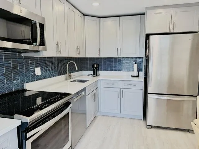 Modern kitchen featuring light gray cabinetry, stainless steel appliances, white countertops, a dark blue subway tile backsplash, and light wood-finish flooring