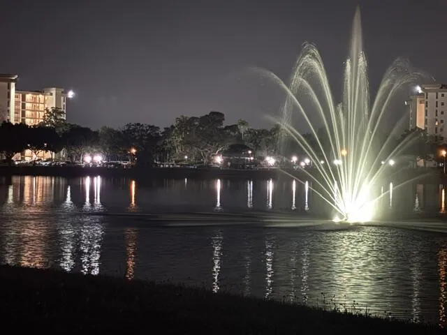 Illuminated water fountain feature centered in a large body of water, with buildings and trees visible on the opposing shore
