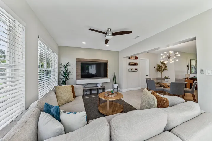Open-concept living area featuring a curved sectional sofa arrangement, wood-finish accent table, ceiling fan with dark metal blades, wall-mounted flat-screen television with built-in media console, recessed lighting, and white shutters
