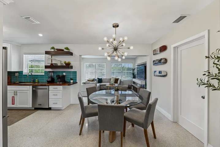 Open-concept dining area with a contemporary chandelier, durable terrazzo flooring, and a kitchen featuring white cabinetry, a teal tile backsplash, and wood-finish countertops