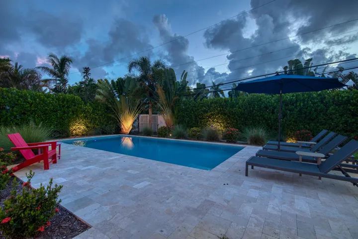 Rectangular swimming pool with light-colored tile surround, surrounded by lush landscaping and uplighting