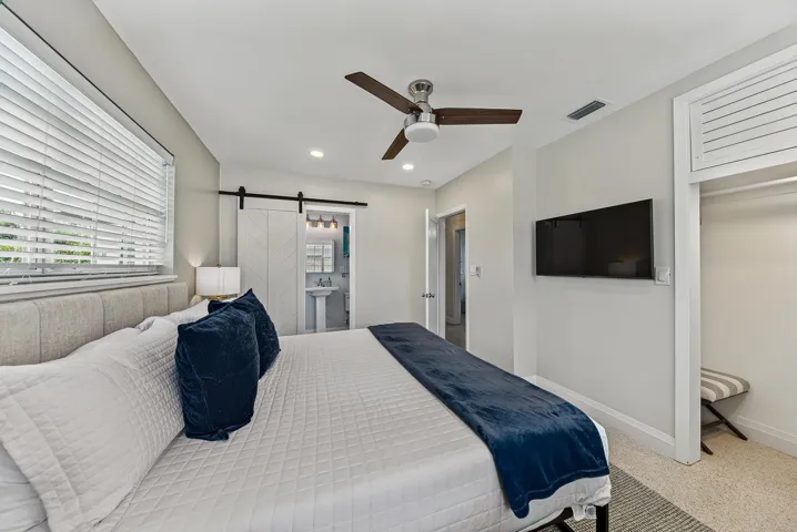 Spacious primary bedroom featuring a wood-finish ceiling fan, recessed lighting, dual windows with plantation shutters, wall-mounted flat-screen television, and direct access to an ensuite bathroom