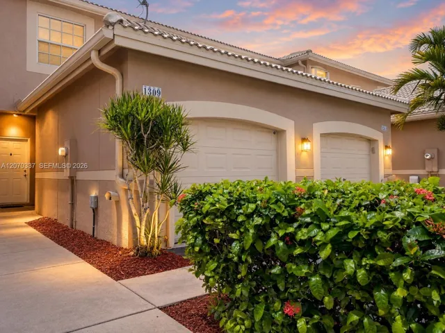 Two-car garage exterior with arched door frames, stucco siding, tiled roof, concrete walkway, and landscaped garden beds