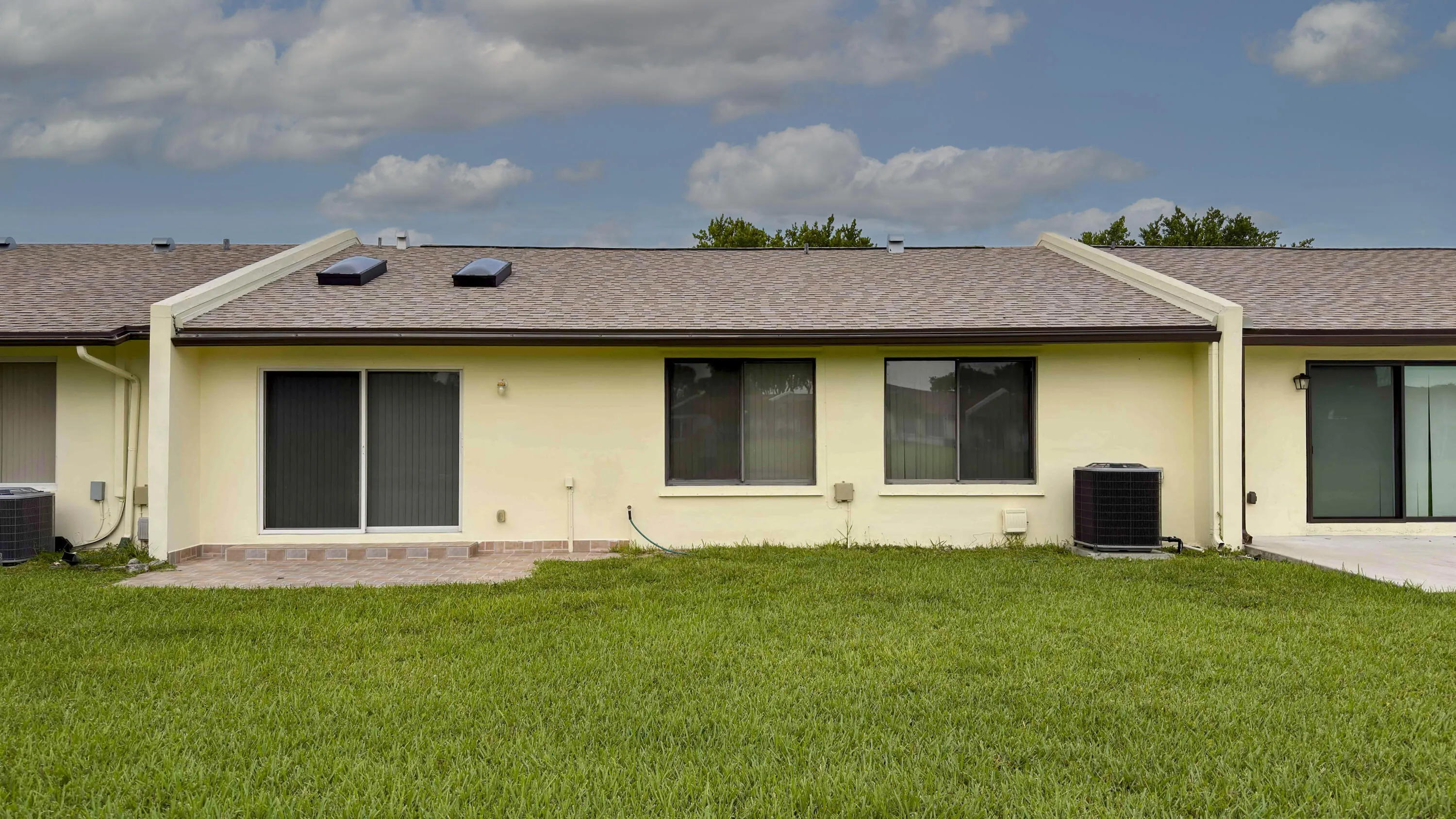 Rear exterior featuring a green lawn, a small paver patio, sliding glass doors, multiple windows, and a shingled roof with skylights