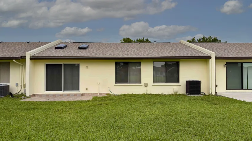 Rear exterior featuring a green lawn, a small paver patio, sliding glass doors, multiple windows, and a shingled roof with skylights