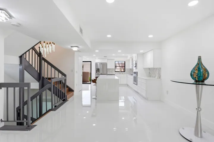 Sophisticated open-concept living space featuring polished light-finish flooring throughout, a curved staircase with metal railings and wood treads, recessed ceiling lighting, and a modern kitchen with white cabinetry and stainless steel appliances visible in the background