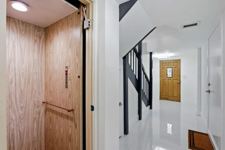 Wood-finish interior elevator with control panel and handrail, alongside a hallway featuring high-gloss flooring, a dark-stained staircase, and a paneled wooden door with a decorative glass insert