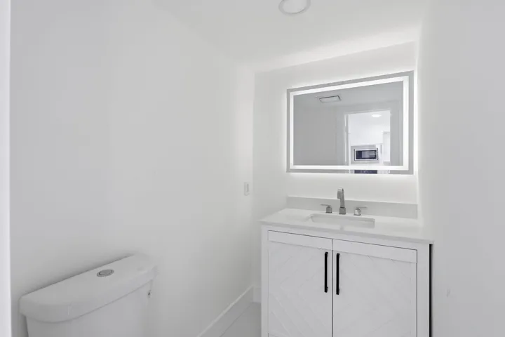 White vanity with textured cabinet doors and dark hardware, integrated rectangular sink, wall-mounted faucet, and illuminated mirror