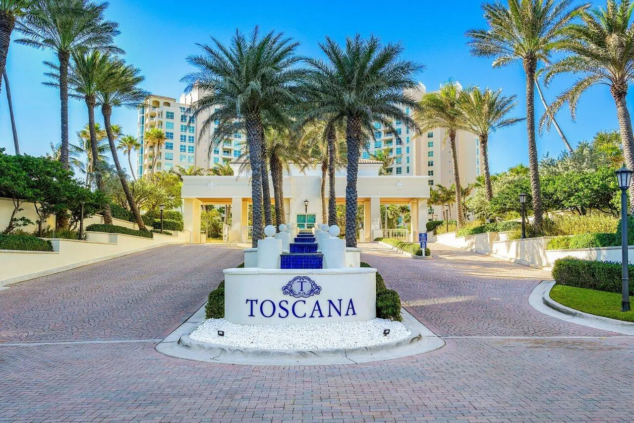 Paver driveway entrance featuring a prominent property sign with a tiered fountain, surrounded by mature palm trees and manicured landscaping