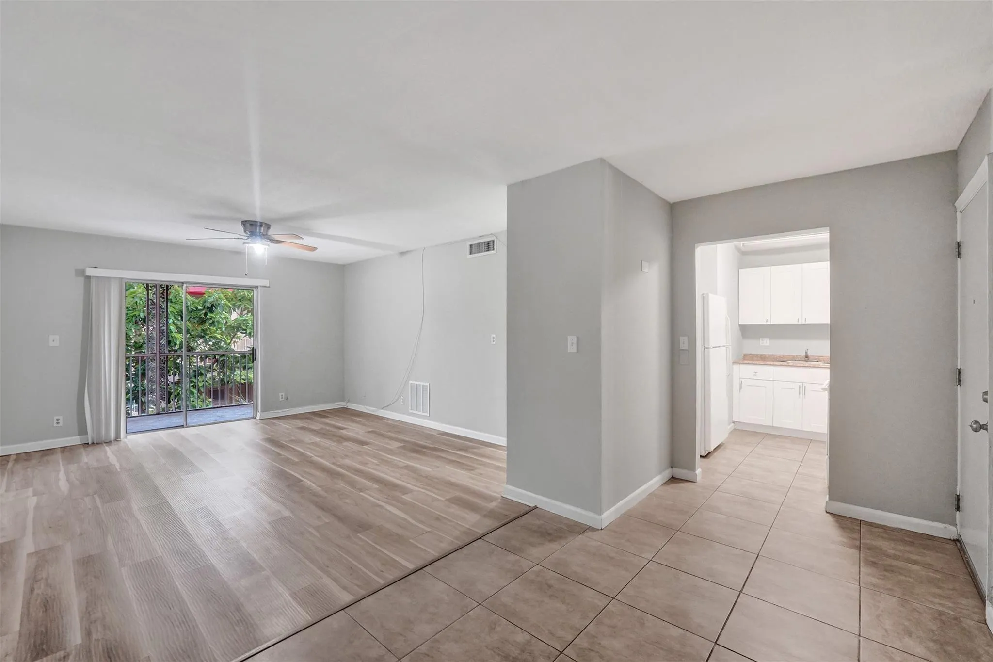 Open-concept living area featuring wood-finish flooring and tile surfaces, abundant natural light from sliding glass doors and multiple windows, neutral gray wall finishes, and a ceiling fan with recessed lighting throughout