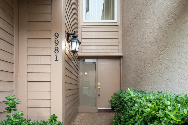 Welcoming entry featuring horizontal siding exterior, illuminated wall sconce, brushed metal door hardware, and lush green landscaping with mature shrubs flanking the entrance