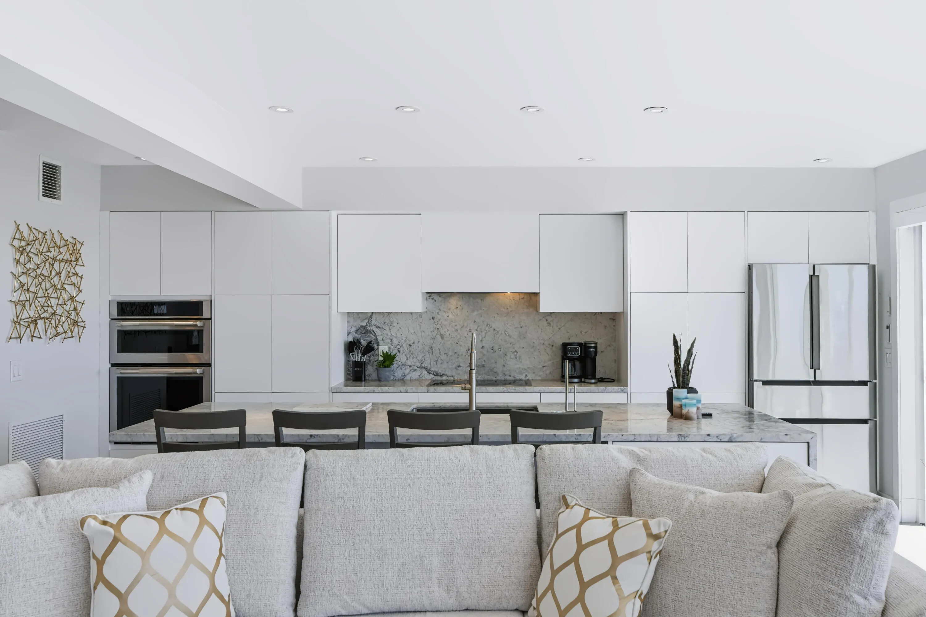 Open-concept kitchen and living area featuring custom white cabinetry, stainless steel appliances including dual wall ovens and a French-door refrigerator, quartz countertops, and a large waterfall-edge island with seating
