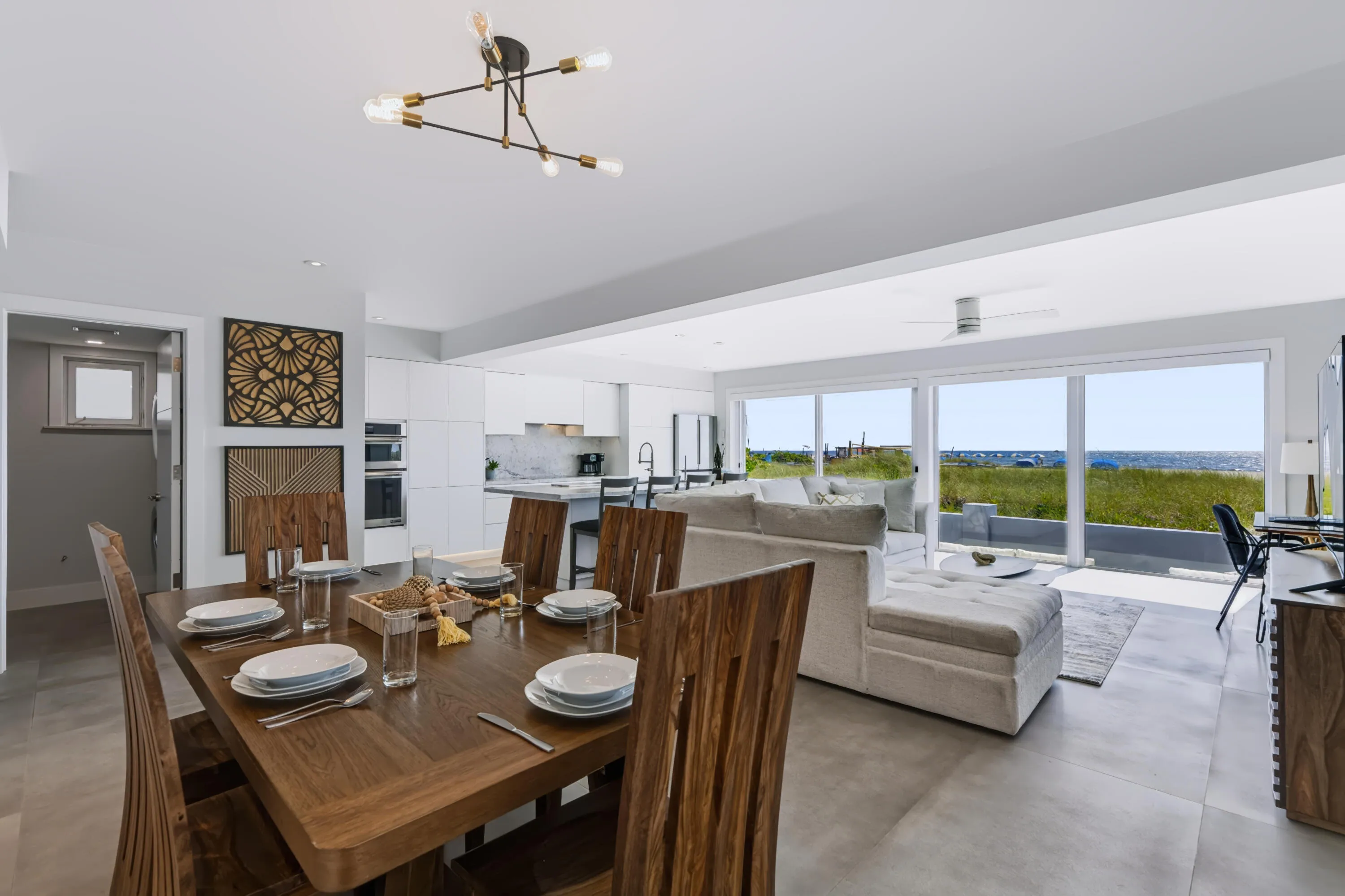Open-concept dining and living area featuring a wood-finish dining table with seating for six, polished concrete flooring, and expansive sliding glass doors overlooking pastoral landscape