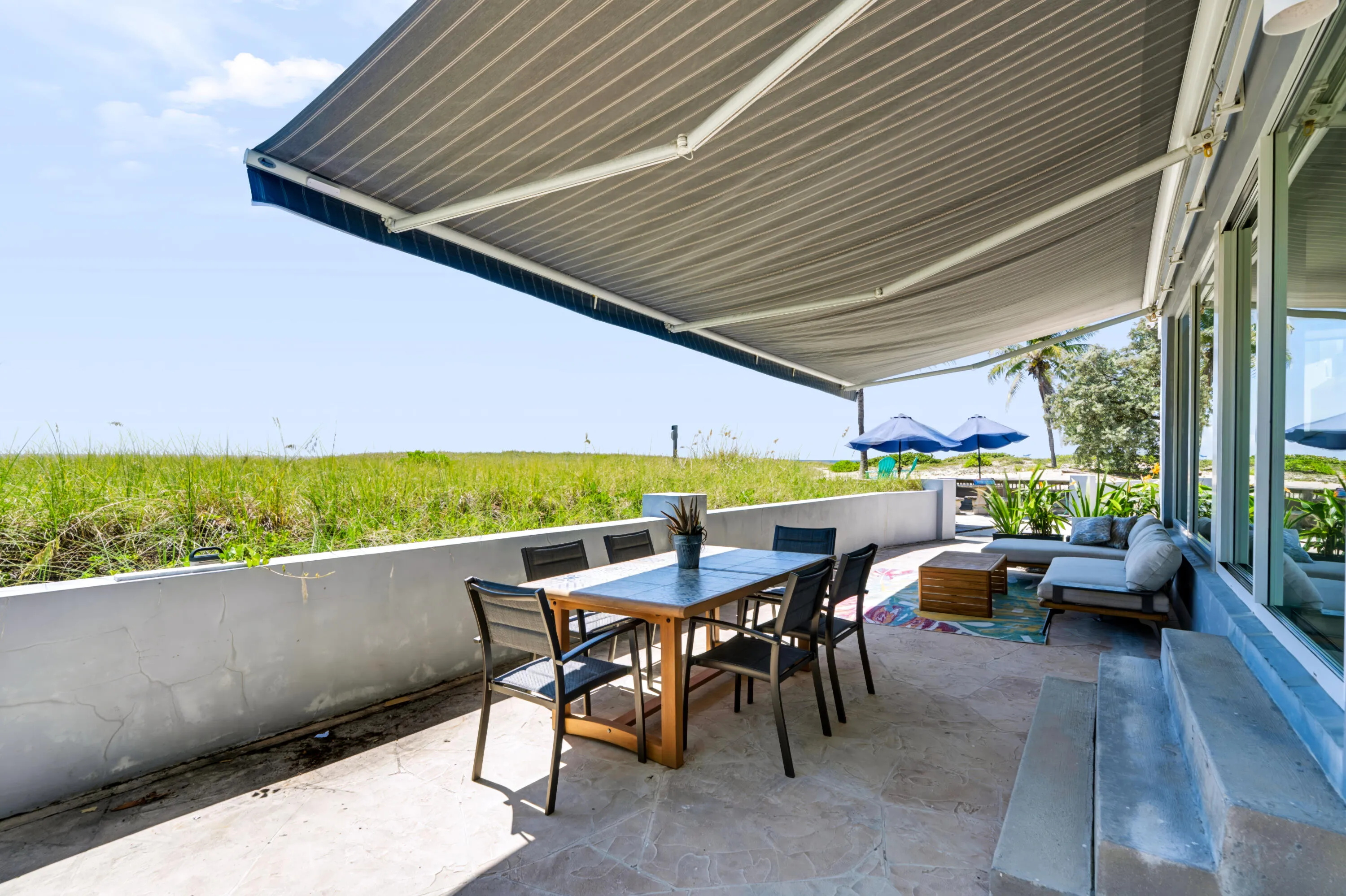 Expansive patio with retractable awning, textured concrete flooring, concrete half-wall, and large glass pane windows