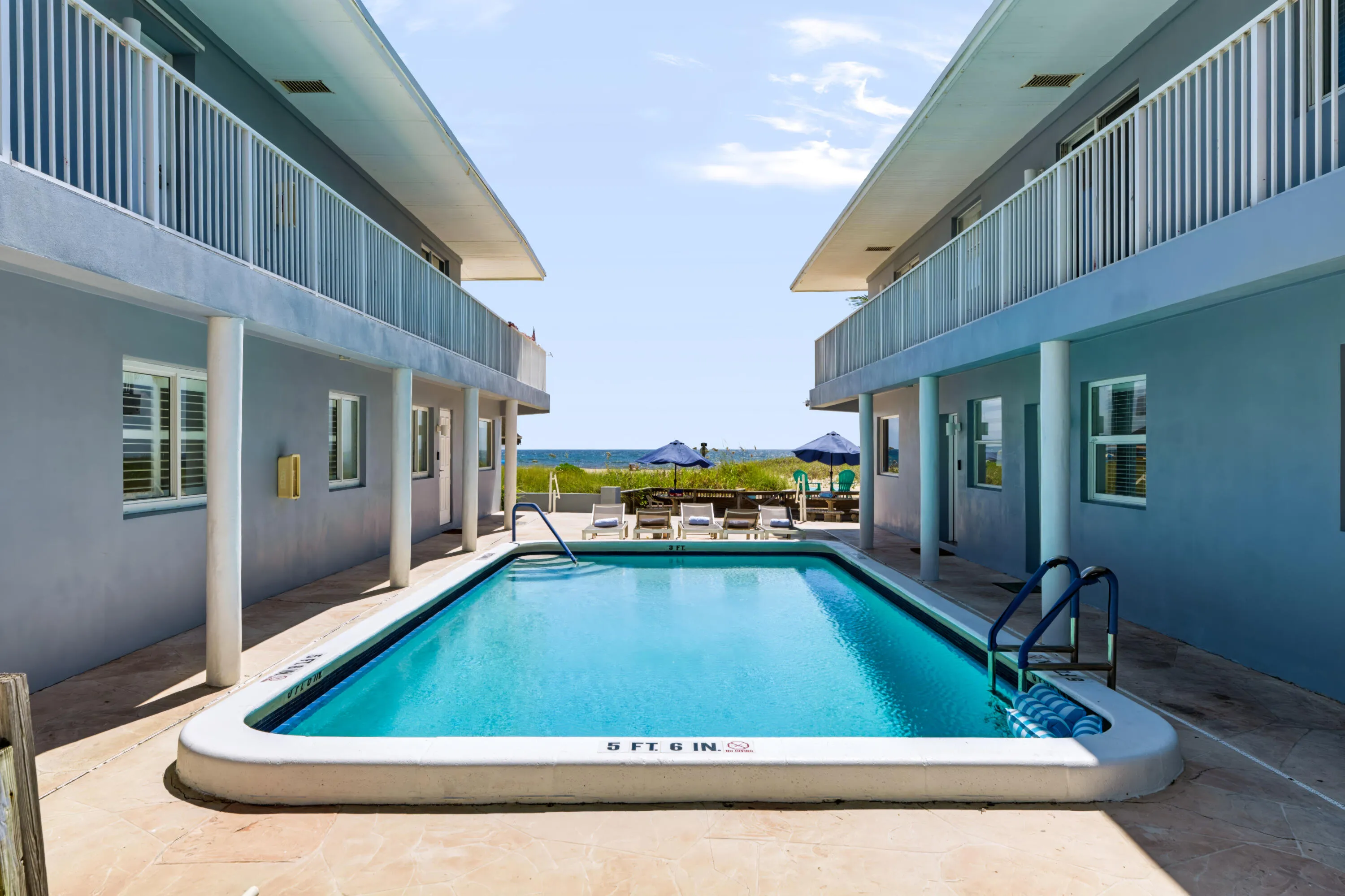 Rectangular swimming pool with clear blue water, surrounded by concrete decking and two-story structures featuring balconies with white railings