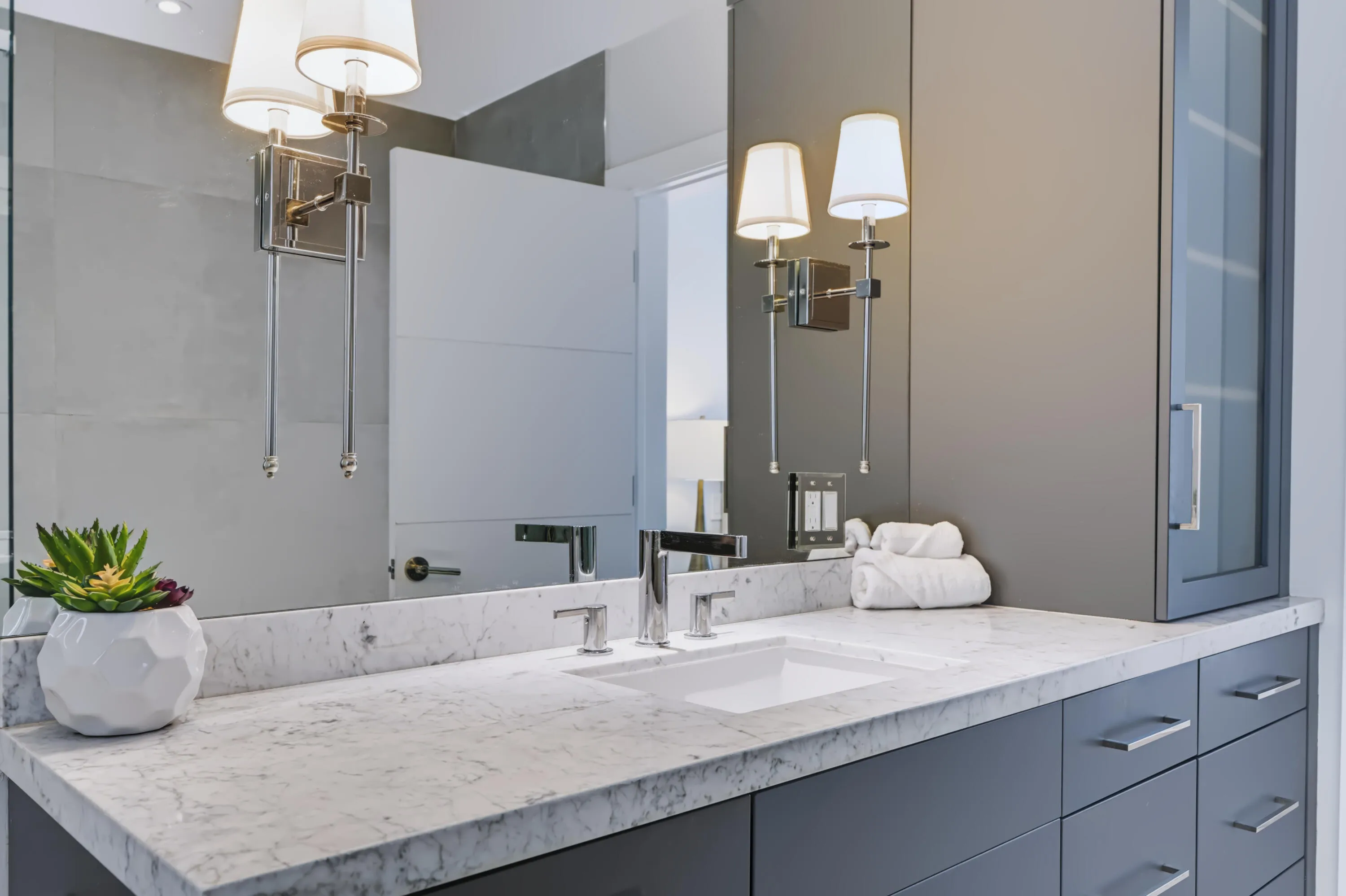 Spa-inspired primary bathroom featuring dual vanities with polished stone countertops, integrated undermount sinks, chrome fixtures, and ample storage cabinetry in soft blue-gray tones