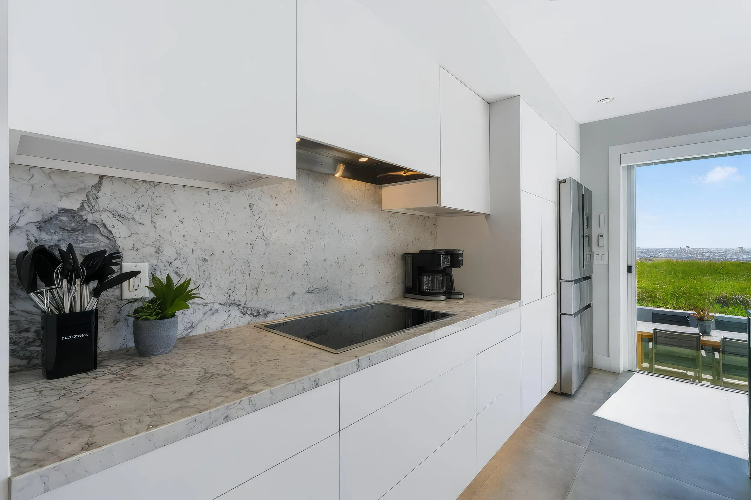 Sleek kitchen featuring white flat-panel cabinetry, integrated cooktop, stainless steel refrigerator, and stone countertops extending to a matching backsplash