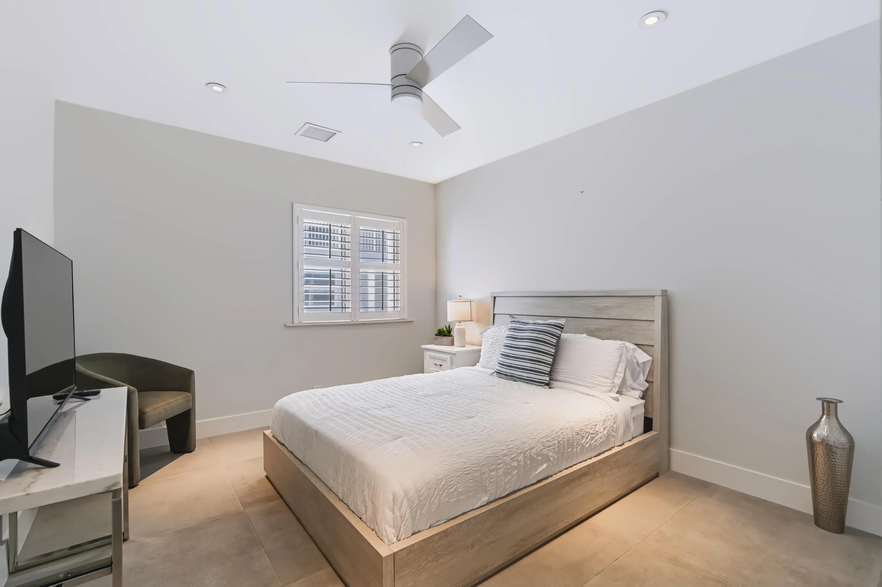 Ceiling fan with integrated lighting, recessed ceiling lights, window with plantation shutters, wood-finish flooring, and a bed frame with a wood-finish headboard