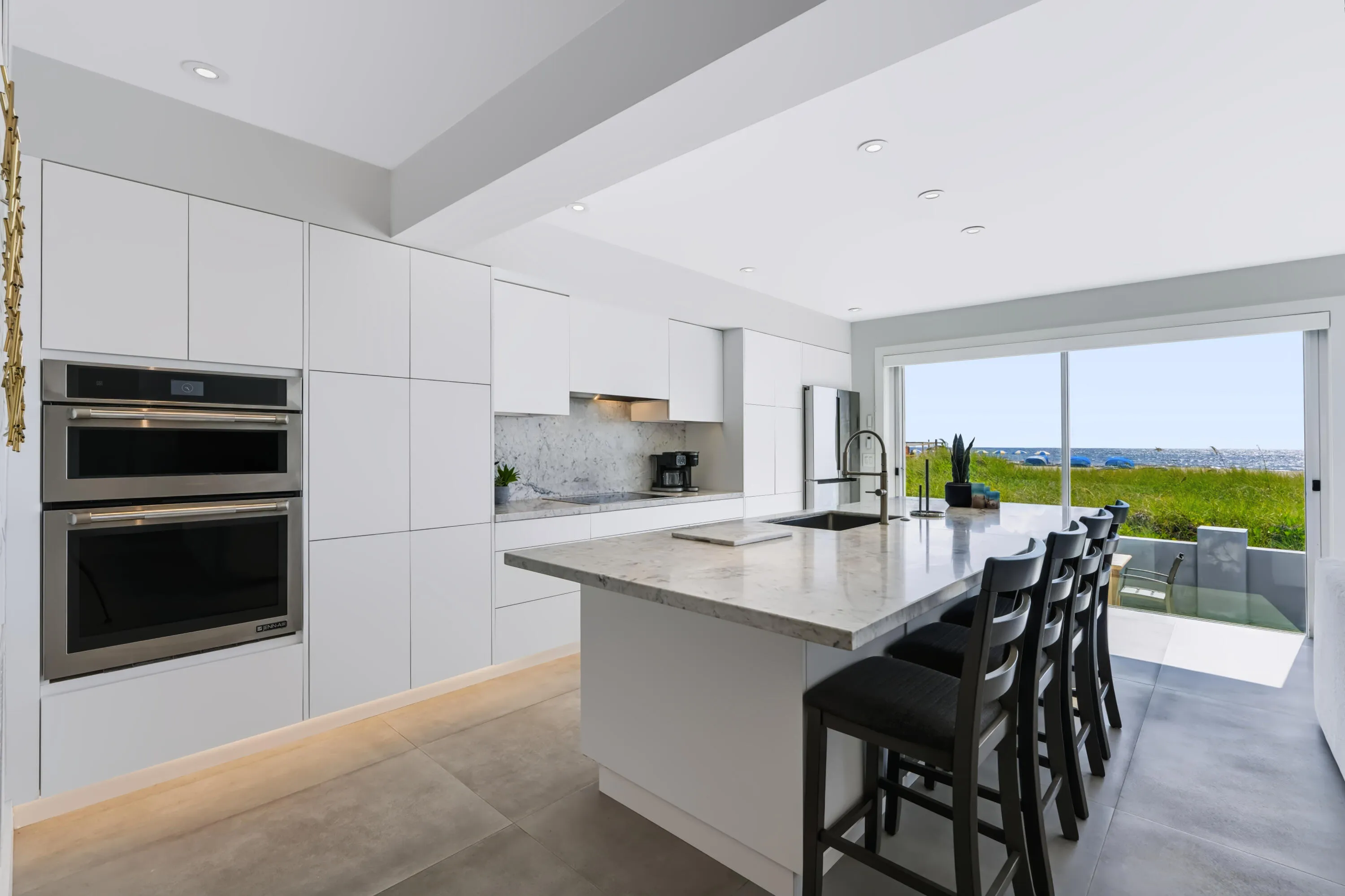 Contemporary kitchen featuring white flat-panel cabinetry, stainless steel double wall ovens, a large island with a built-in sink, and a sliding glass door leading to a patio
