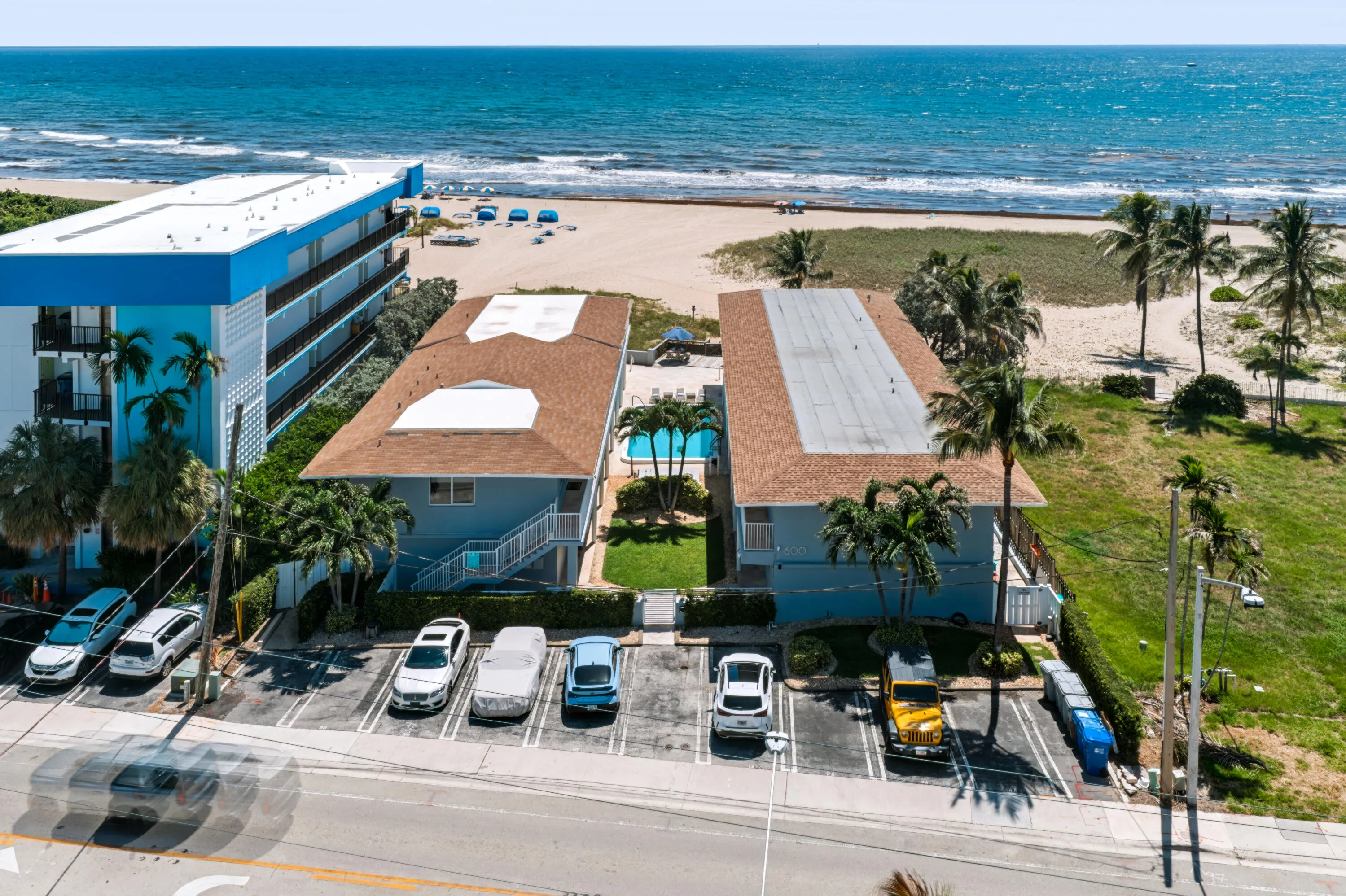 Beachfront property with direct access to a sandy shoreline, featuring a swimming pool, mature palm trees, and an asphalt parking area