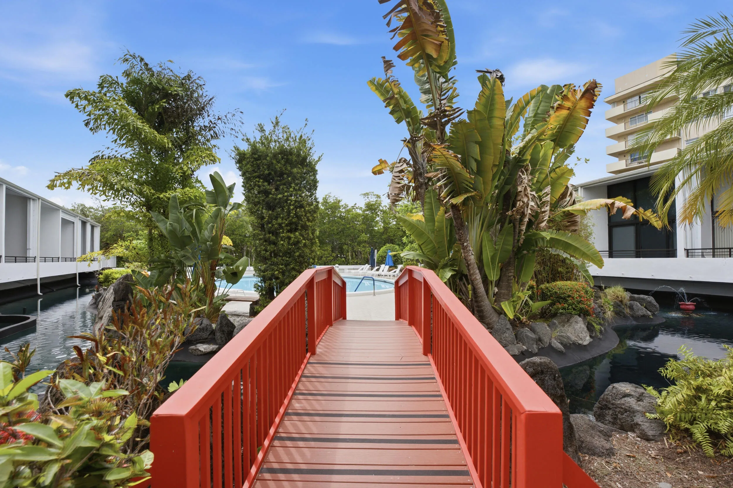 Red wooden pedestrian bridge over a dark water feature, surrounded by dense tropical foliage and rock landscaping