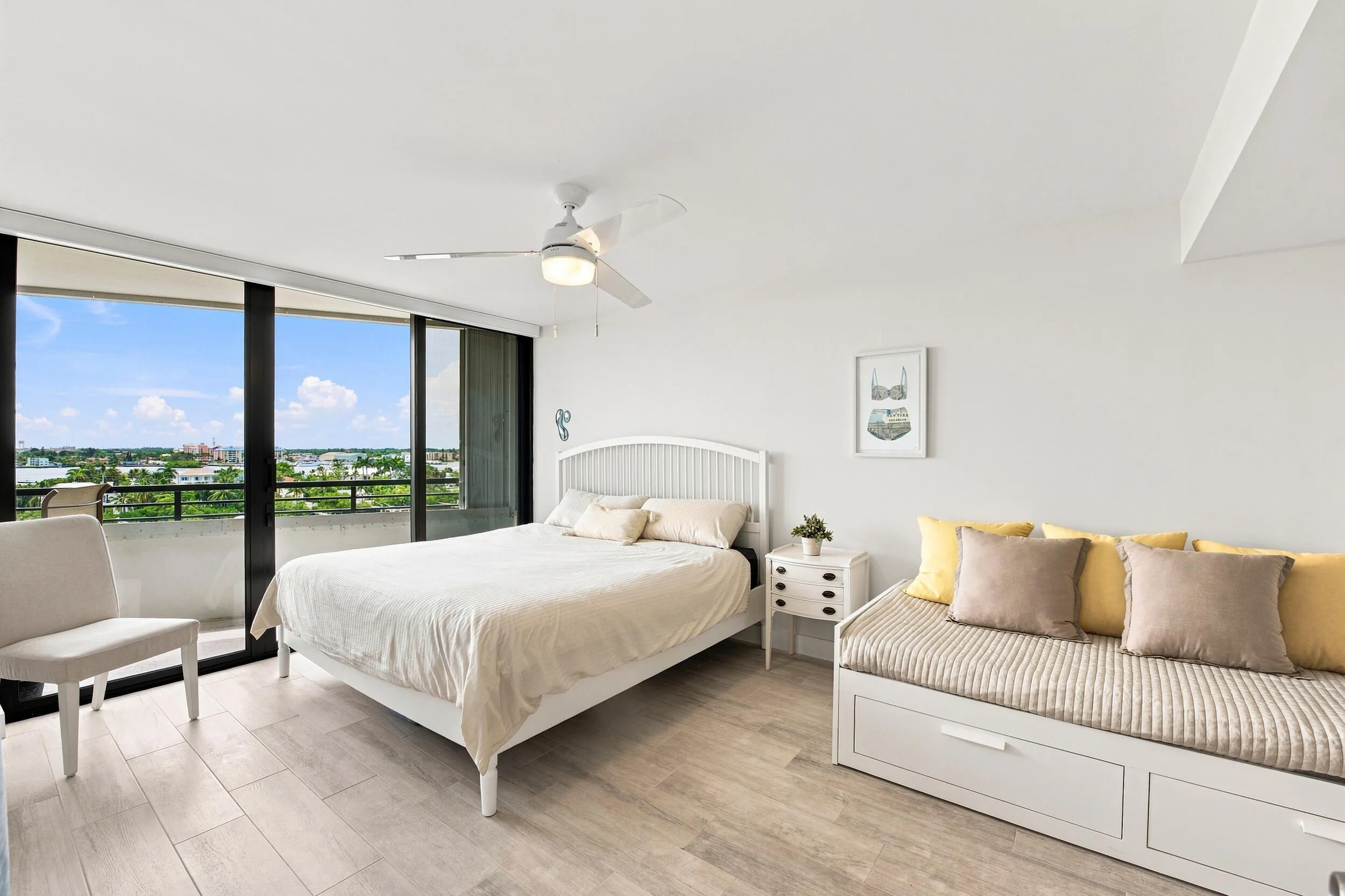 Expansive windows frame a body of water and distant cityscape, complemented by wood-finish flooring, a ceiling fan with integrated lighting, and a daybed with built-in storage drawers