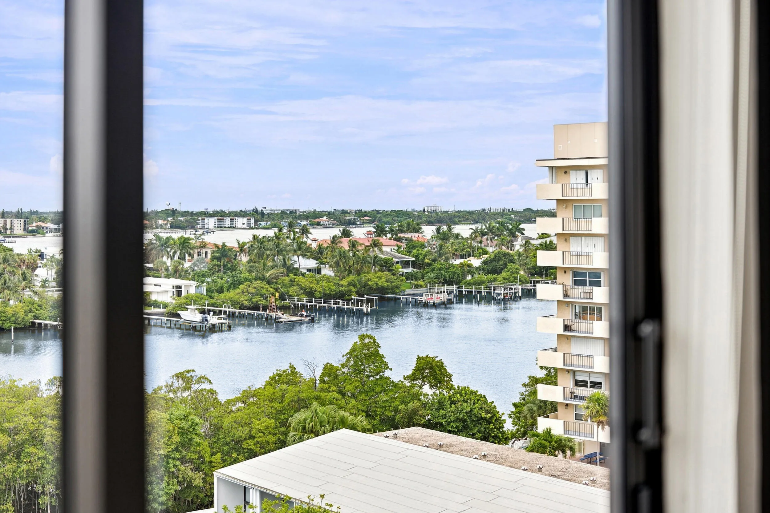 Expansive water vista framed by an architectural window, featuring a waterway, distant residential properties, lush green landscaping, and a multi-story building with multiple balconies