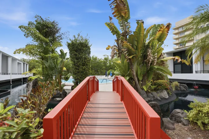 Red wooden pedestrian bridge over a dark water feature, surrounded by dense tropical foliage and rock landscaping