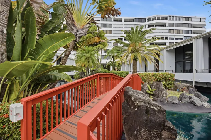 Decorative red pedestrian bridge over a water feature, surrounded by tropical landscaping, with a multi-story building in the background