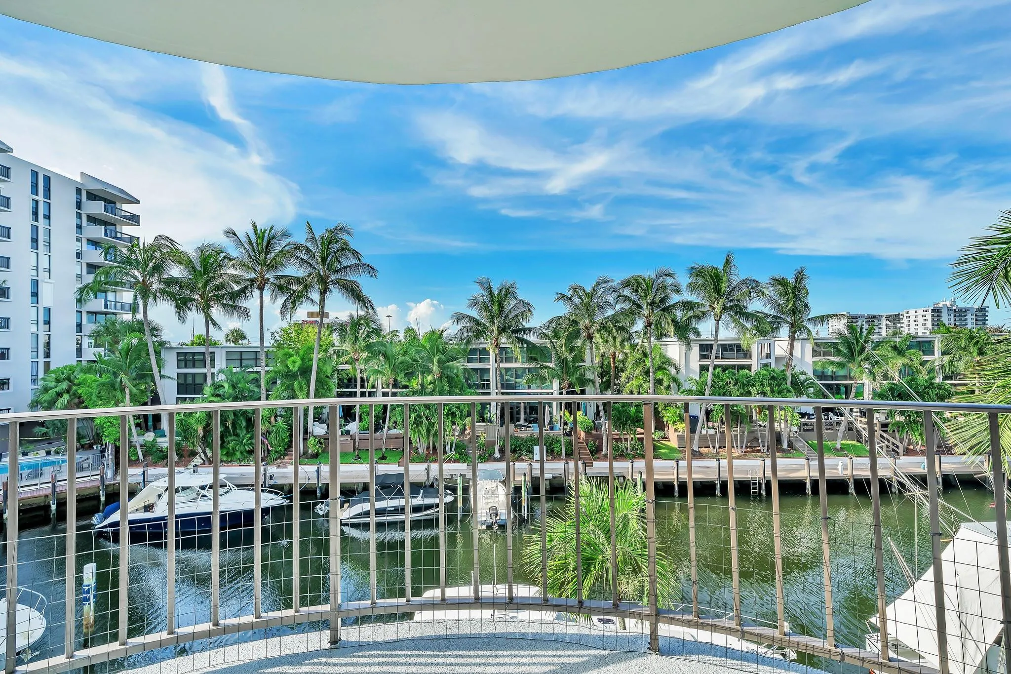 Curved balcony with metal railing overlooking a waterway with boats, palm trees, and residential buildings
