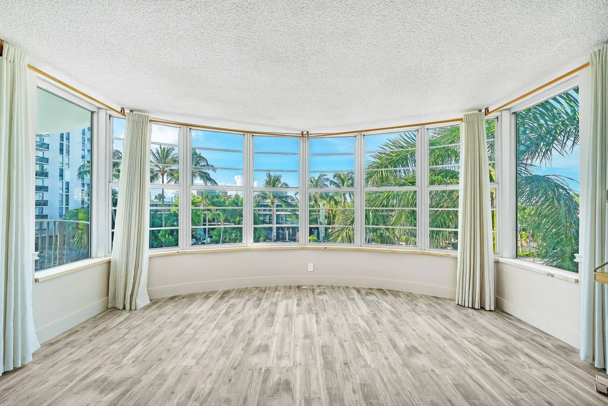 Curved wall of windows, light wood-finish flooring, white window frames, tropical scenery, and distant water