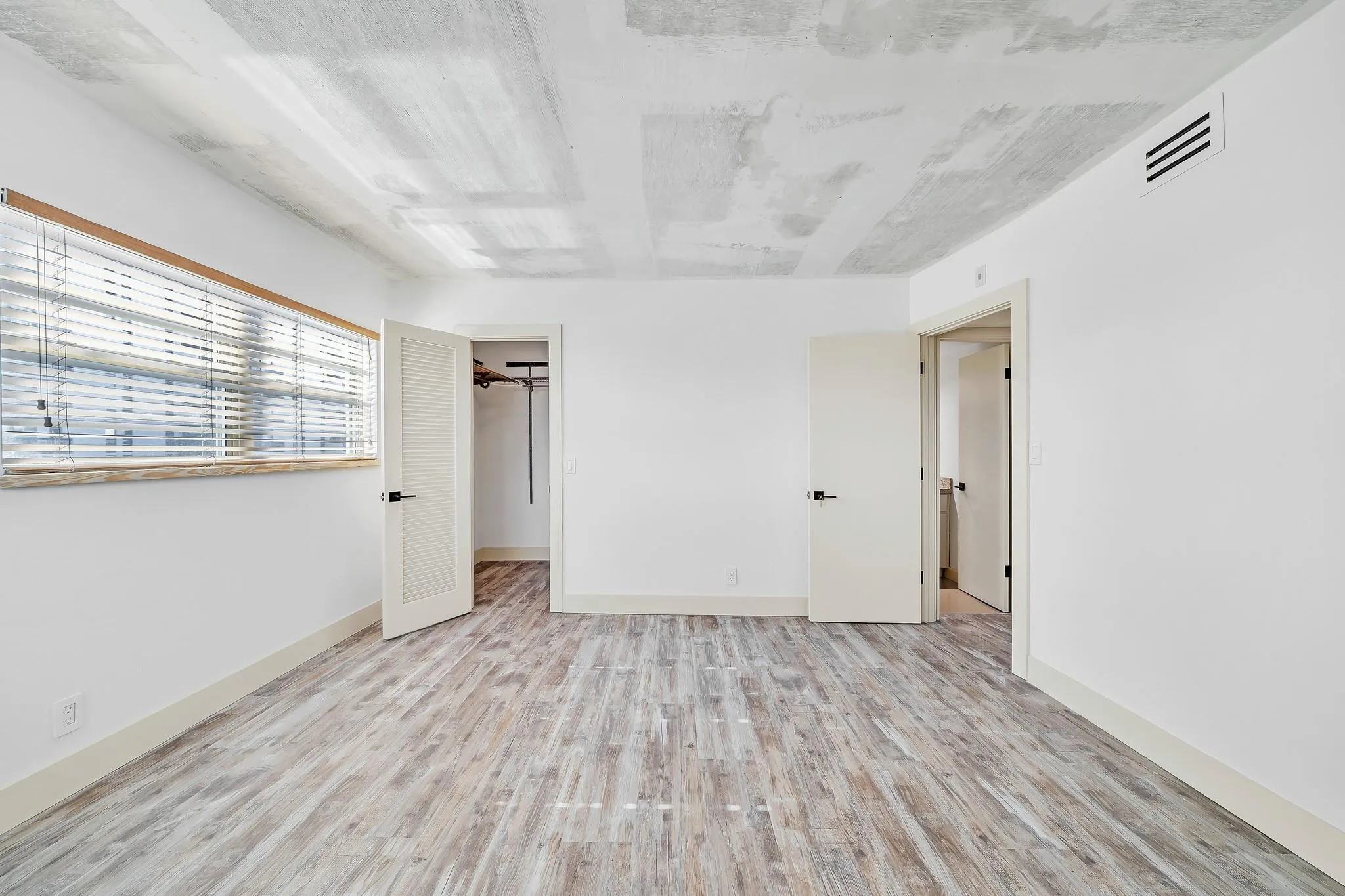 Wood-finish flooring, white walls, louvered closet door, window with horizontal blinds, and exposed concrete ceiling