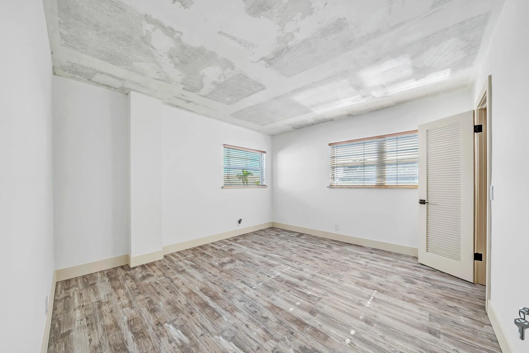 Concrete ceiling, wood-finish flooring, two windows with blinds, louvered door, white walls