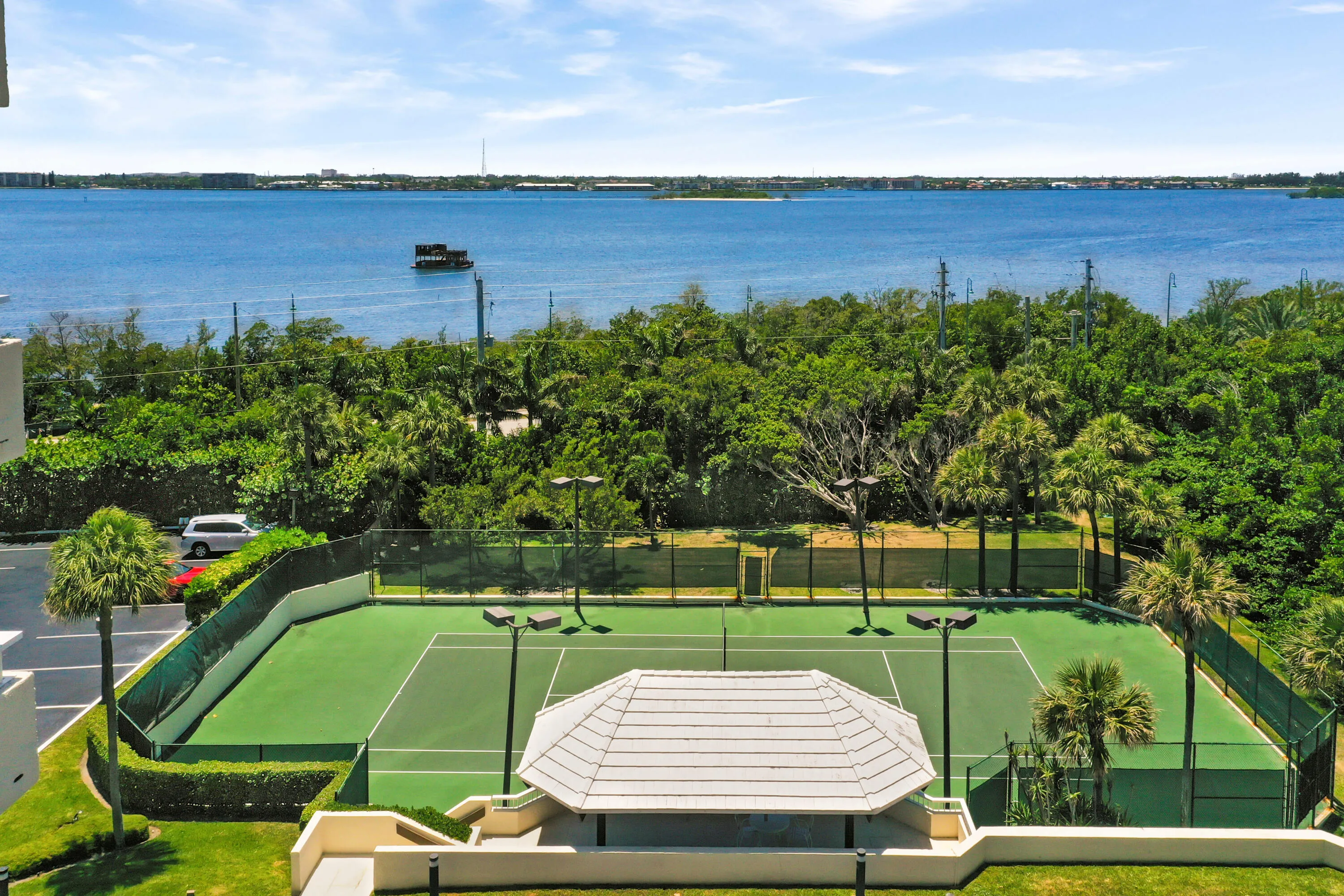 Private tennis court with a shaded seating structure, surrounded by lush tropical landscaping and a body of water