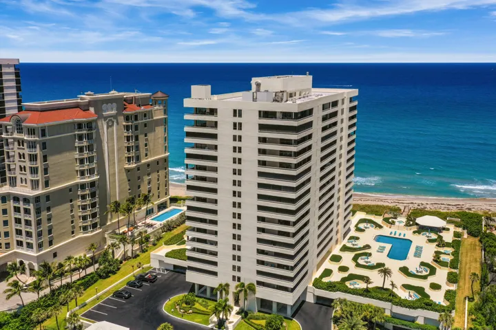 Oceanfront high-rise building with multiple balconies, a ground-level swimming pool, and an adjacent parking area