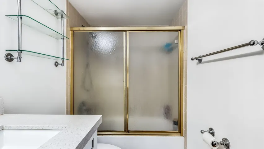 Shower with sliding frosted glass doors and gold-tone trim, light tile surround, built-in glass shelving, and a white countertop with integrated sink
