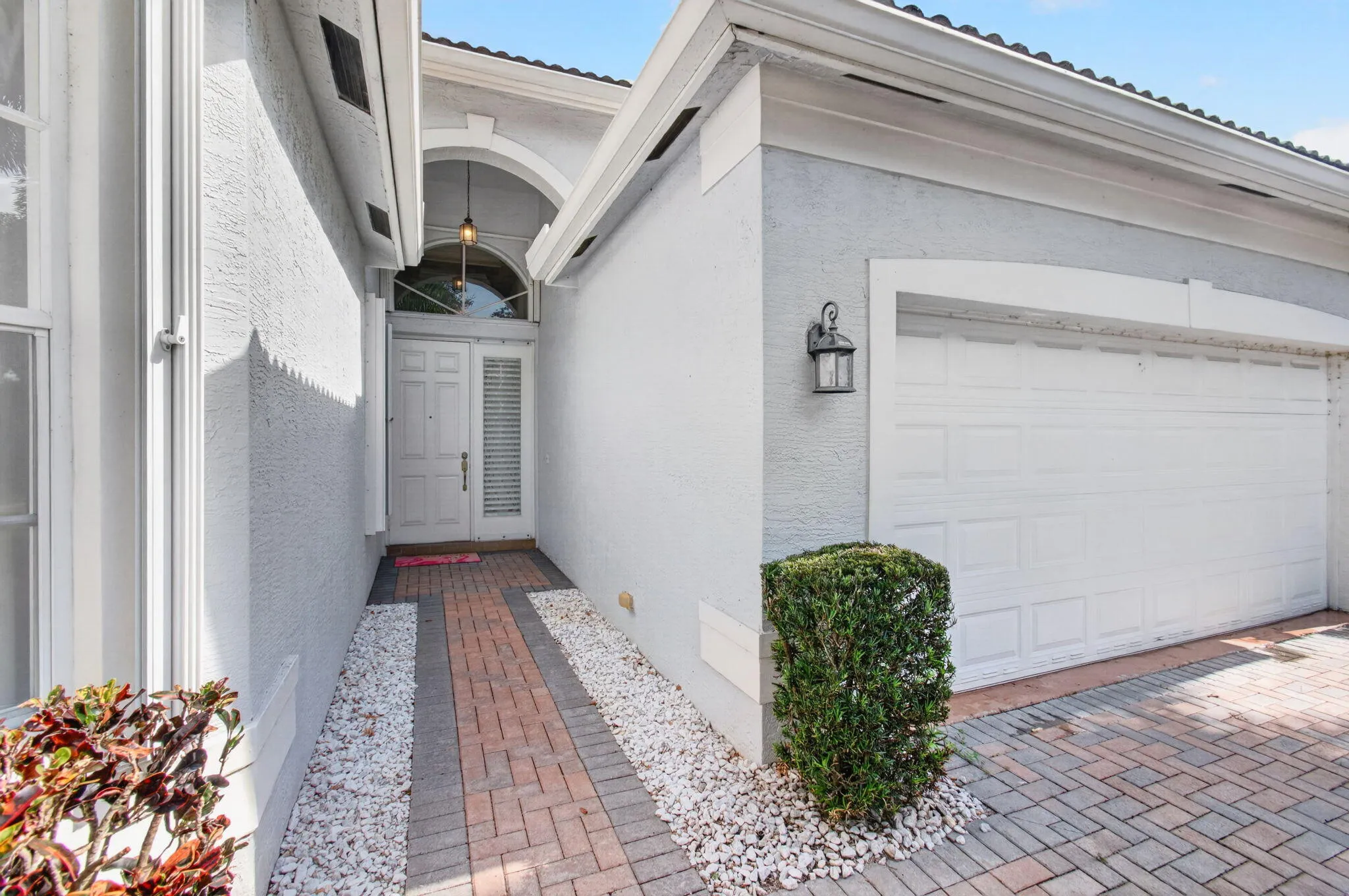 Entryway with arched transom window, brick paver walkway, white gravel landscaping, and attached garage with exterior light fixture