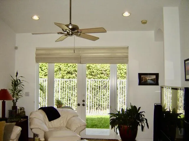 Bright, light-filled living space featuring French doors with vertical blinds opening to outdoor grounds, ceiling fan with brass finish, recessed lighting throughout, and abundant natural light from multiple glass panels