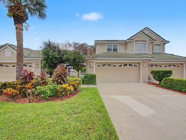 Two-story residence featuring a three-car garage with decorative door panels, complemented by mature landscaping including tropical palms, flowering shrubs with deep burgundy and golden foliage, and manicured lawn fronting the property