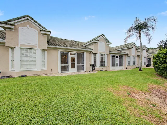 Expansive rear grounds featuring manicured lawn and mature tropical landscaping, anchored by a multi-section residence with arched architectural details, dual French doors, and large windows framing the outdoor space