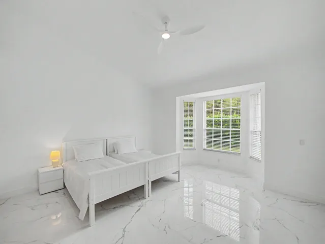Bright secondary bedroom featuring polished stone-finish flooring, white painted wood bed frame, ceiling fan with integrated lighting, and French doors opening to natural light and outdoor access via plantation shutters