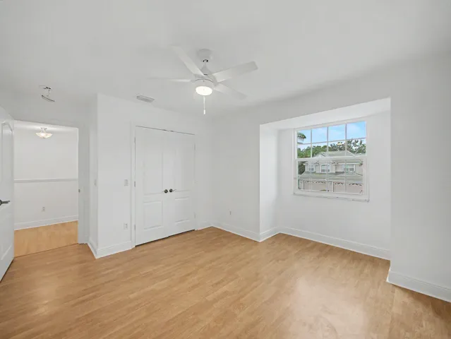 Spacious bedroom featuring wood-finish flooring, a ceiling fan with integrated lighting, generous natural light from a large window, and ample closet storage with double doors