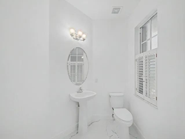 Bright powder room featuring a pedestal sink, oval mirror with decorative frame, chrome wall-mounted sconce lighting, and toilet with white ceramic fixtures