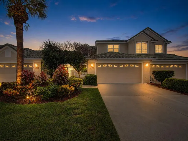 Elegant two-story residence illuminated by professional landscape lighting, featuring a triple-car garage with decorative hardware, manicured lawn with mature palm and tropical plantings, and architectural details including upper-level windows and neutral exterior finishes