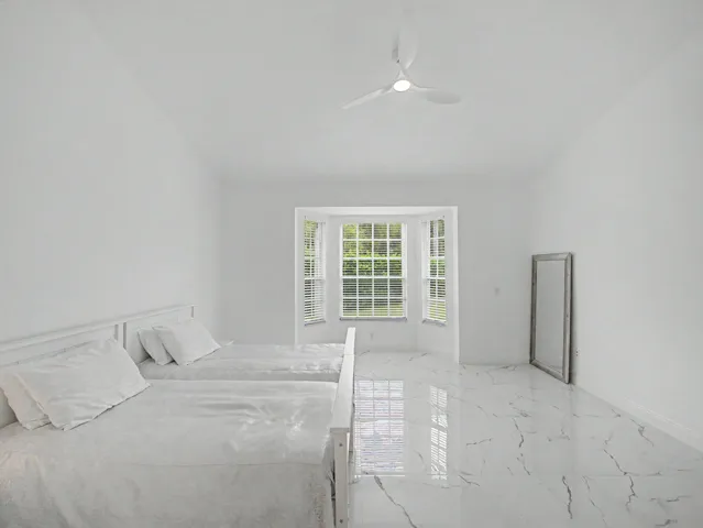 Bright, light-filled bedroom featuring polished stone flooring with linear detailing, large multi-pane French doors with transom windows, ceiling fan, and clean white wall finishes throughout