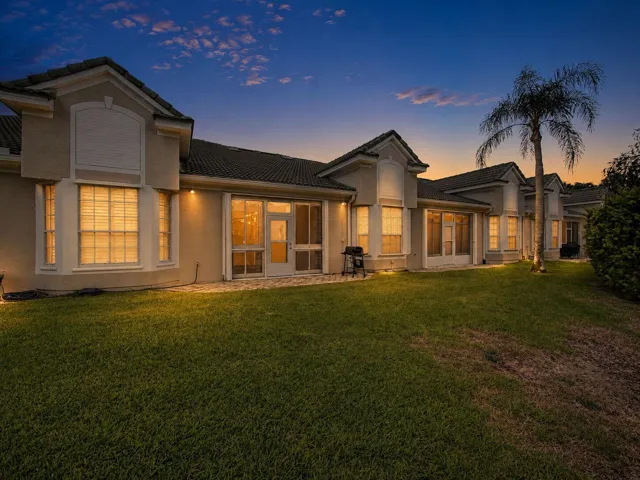 Elegant residence featuring a stucco exterior with arched architectural details, expansive lawn with mature landscaping including palm trees, multiple sets of French doors and large windows with interior lighting, and a covered patio area ideal for outdoor entertaining