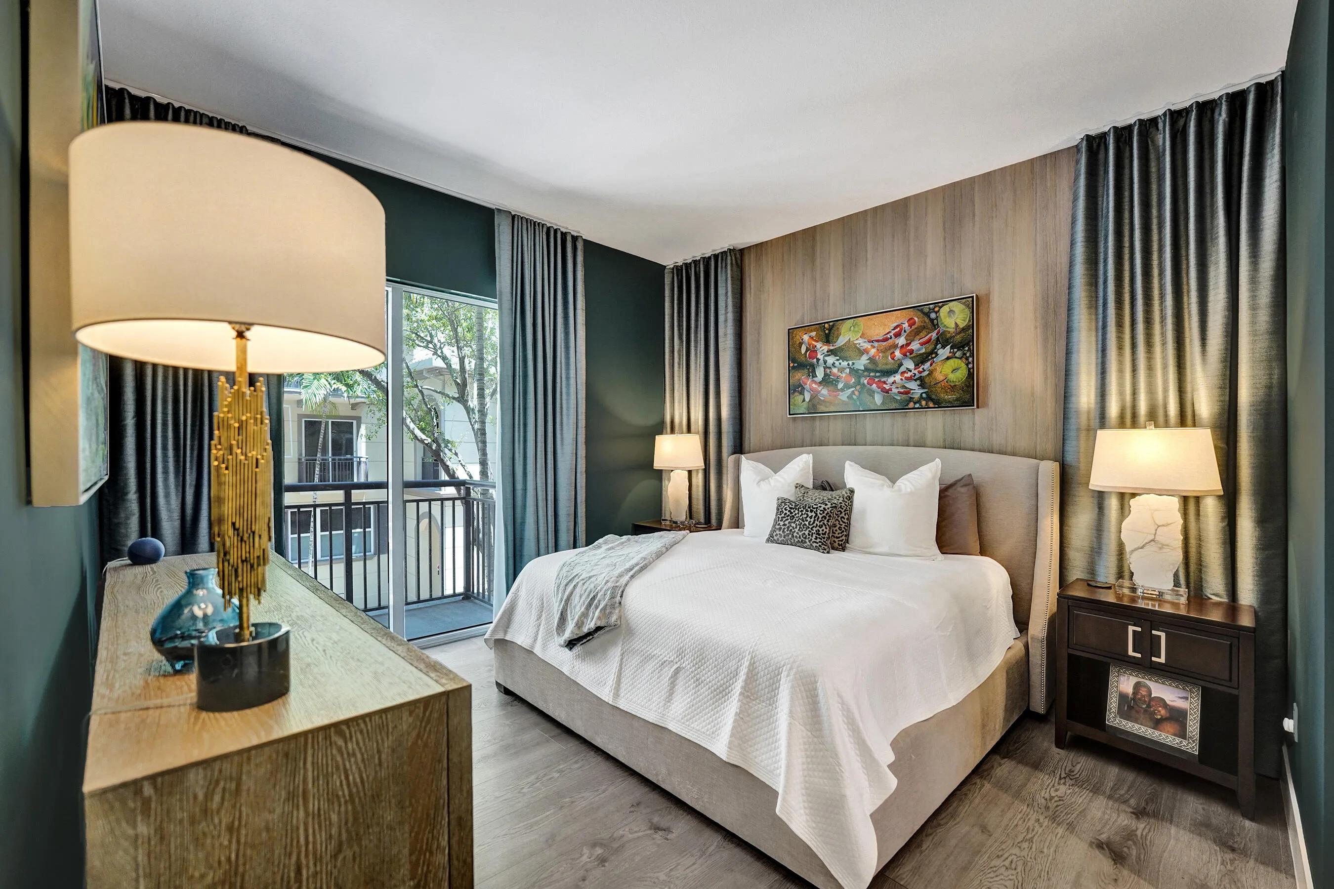 Wood-finish flooring, accent wall with vertical wood paneling, full-height drapery, balcony access, and built-in bedside lighting