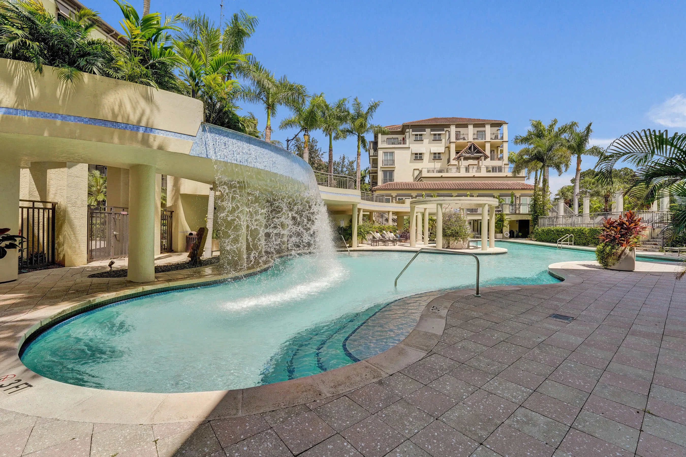 Resort-style pool with a waterfall feature, surrounded by paver decking and lush tropical landscaping